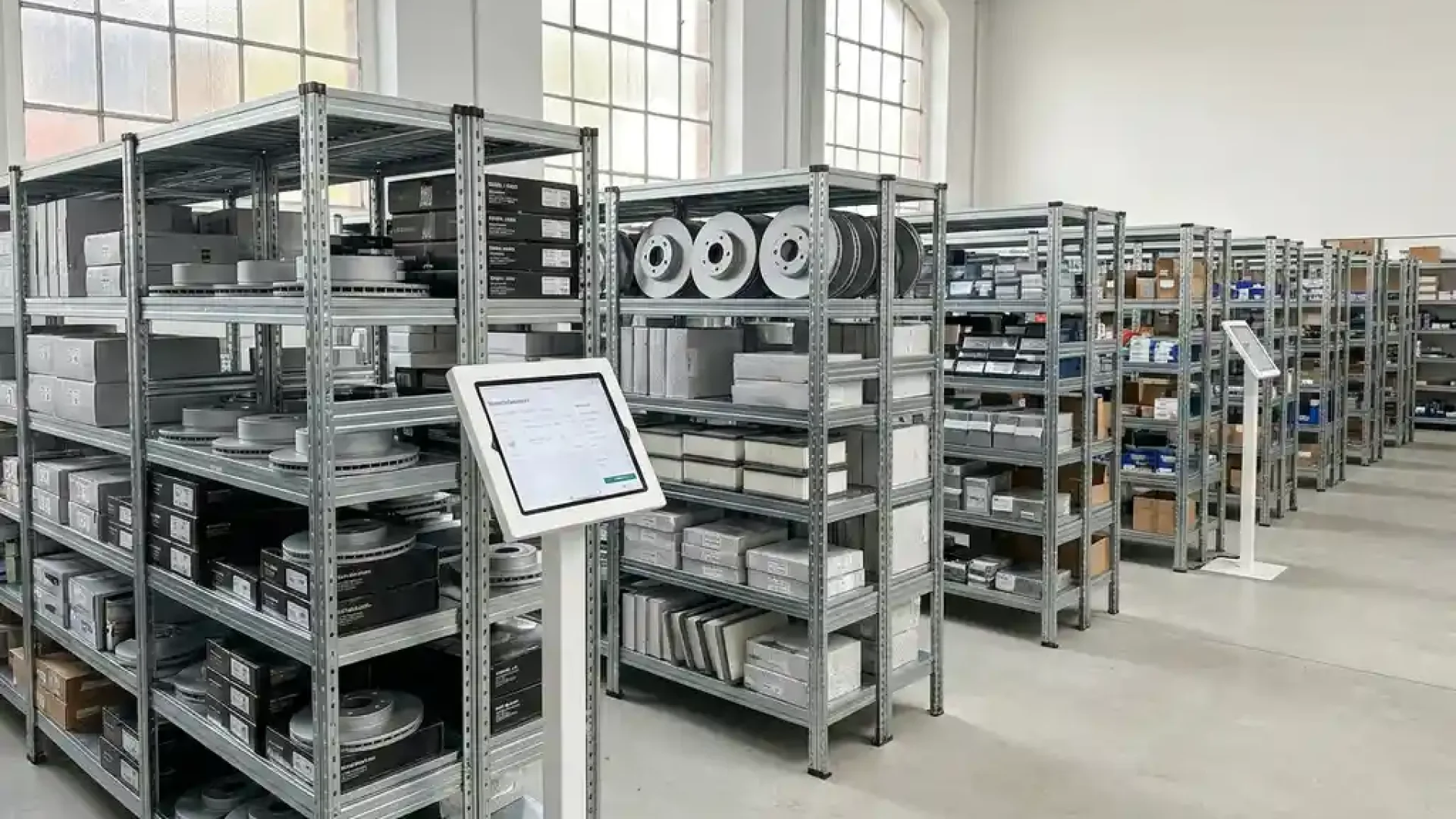 Ordentliches Kfz-Ersatzteillager in Deutschland mit Bremsscheiben auf Metallregalen und einem Tablet mit B2B-Bestelloberfläche.