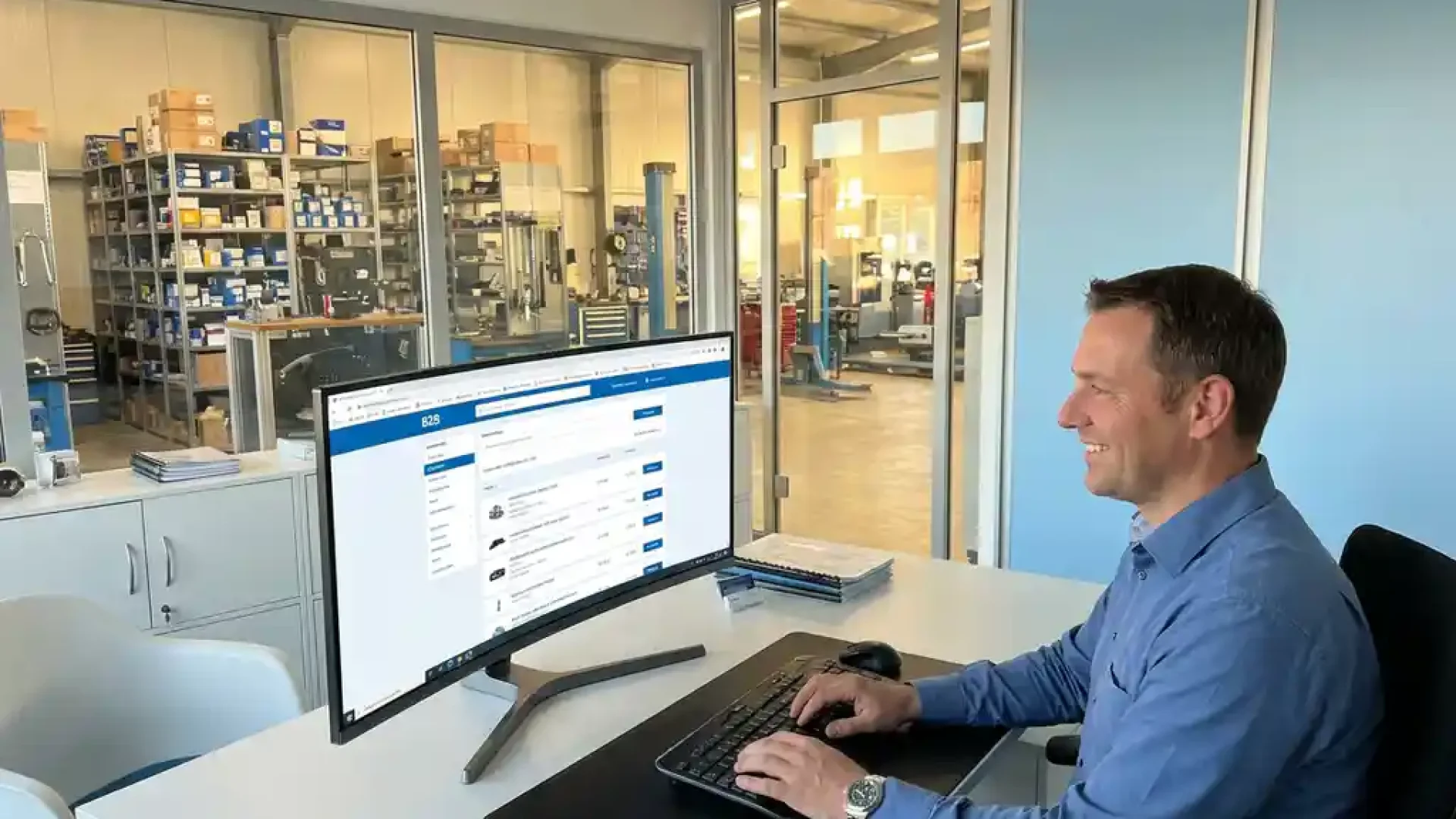 Kfz-Teile-Händler am Monitor beim Durchsuchen eines B2B-Webshops in modernem deutschen Werkstattbüro mit Ersatzteilen im Regal.
