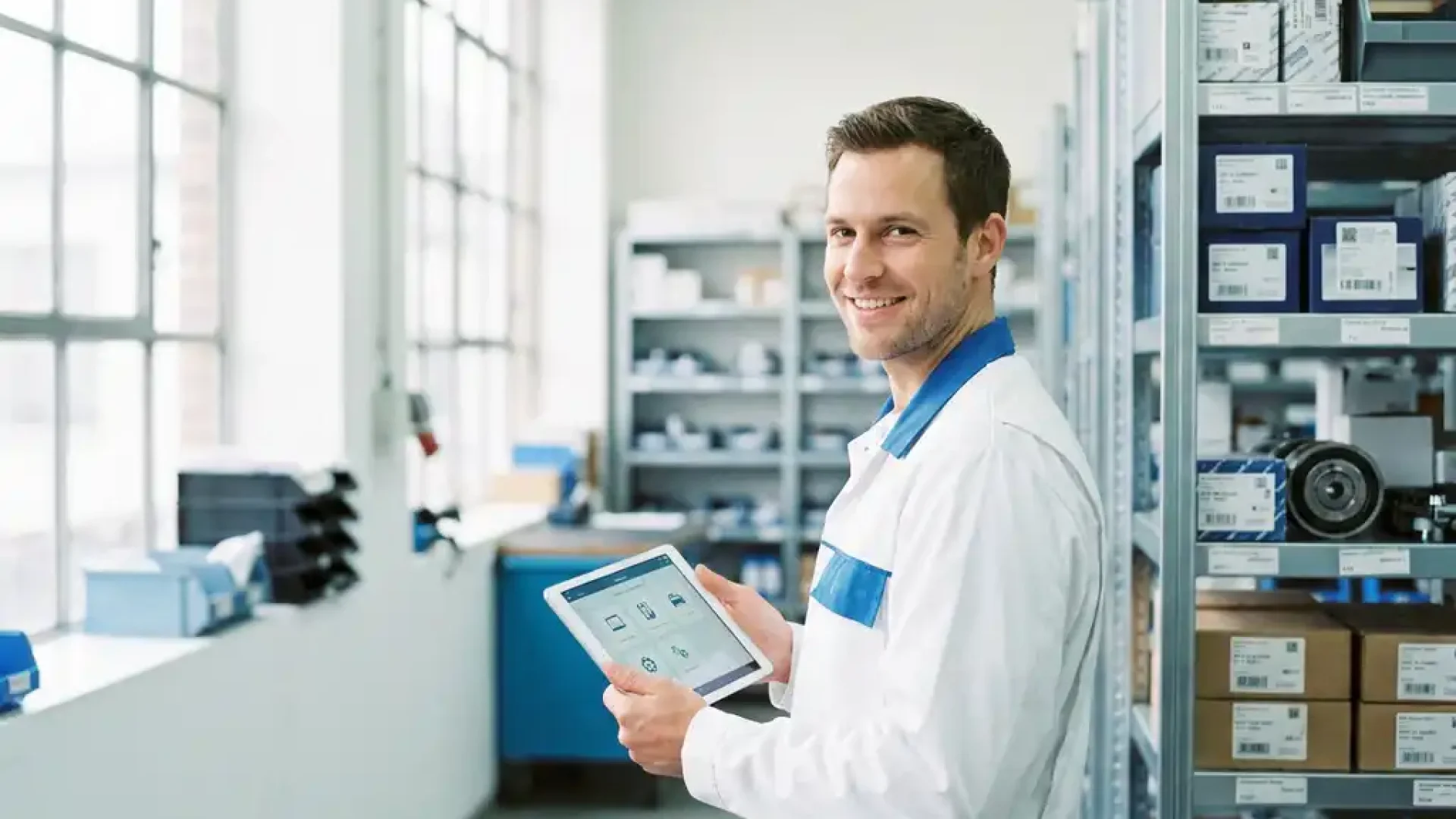 Mechaniker in moderner Kfz-Werkstatt bedient Tablet mit Ersatzteil-Software, sortierte Regale im Hintergrund.